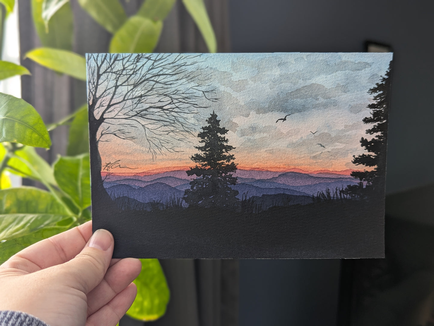 Hand holding a painting of a sunset over mountains with trees.