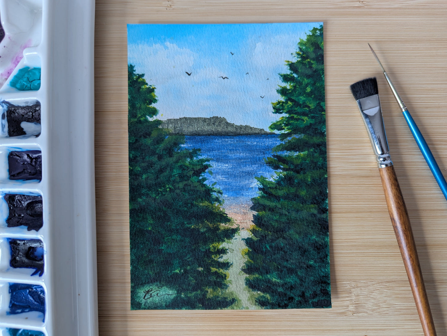 Painting of a landscape with trees and water, placed on a wooden surface with art supplies.