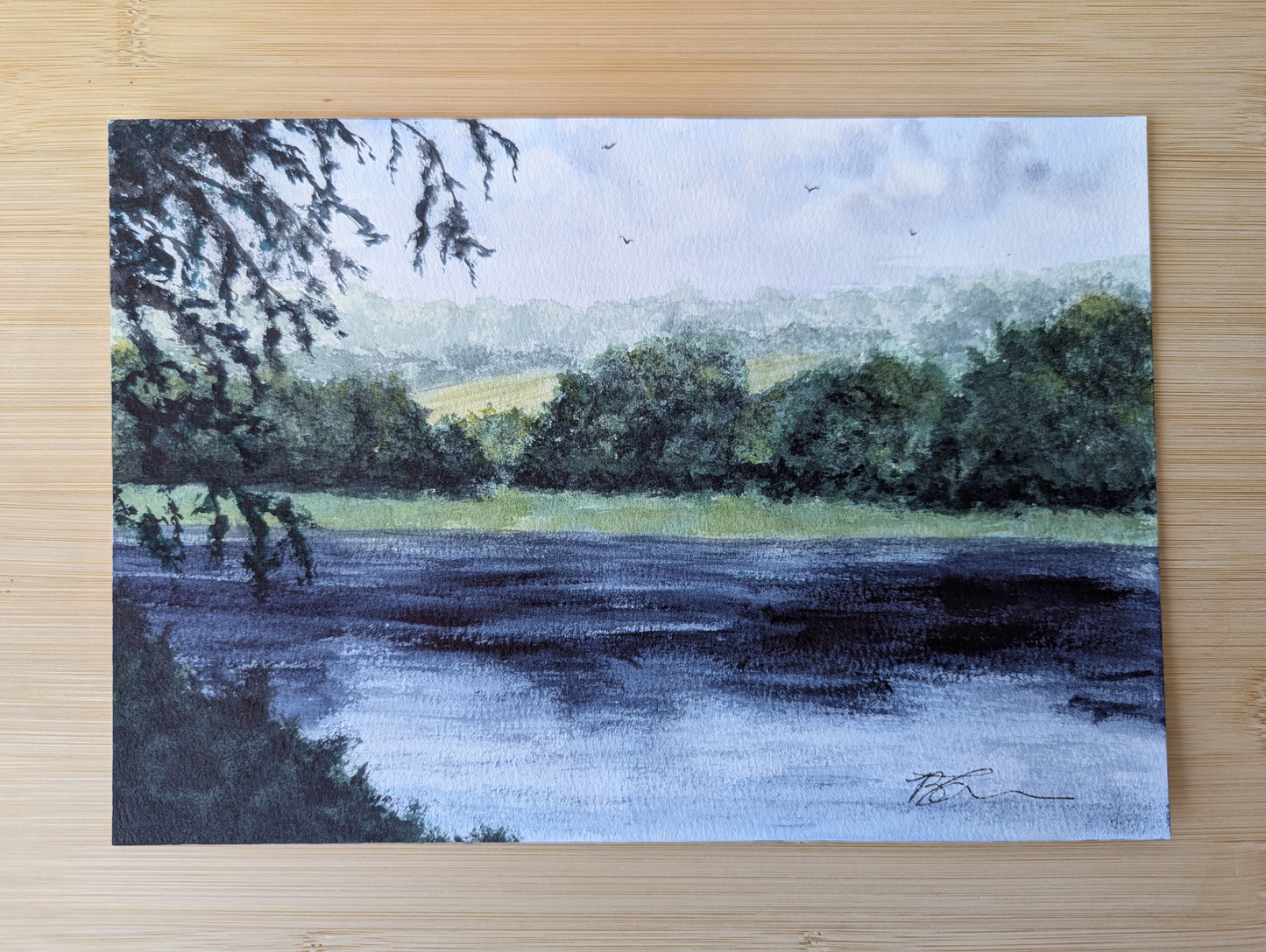 Watercolour Painting of a landscape with trees and water laying flat on a wooden surface