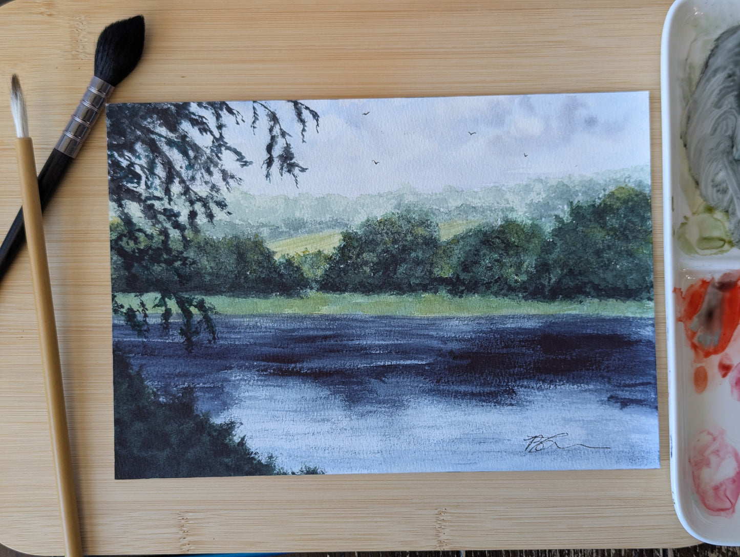 Painting of a landscape with dark sky and green fields on a wooden surface with art supplies.