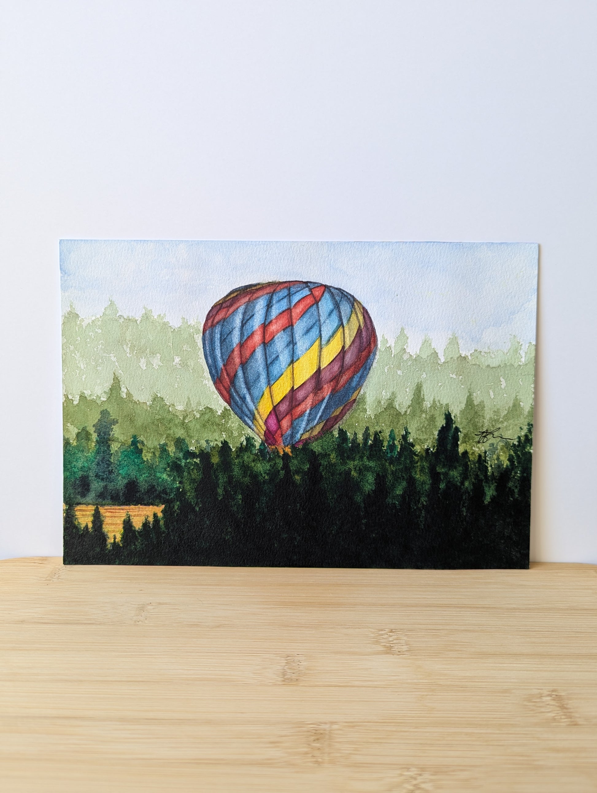 Watercolour painting of colourful hot air balloon taking off from afield surrounded by pine trees in the morning. Painting is sitting on a light wood table and leaning against a white wall 