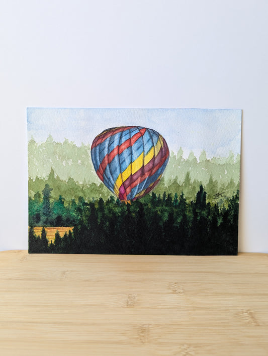 Watercolour painting of colourful hot air balloon taking off from afield surrounded by pine trees in the morning. Painting is sitting on a light wood table and leaning against a white wall 