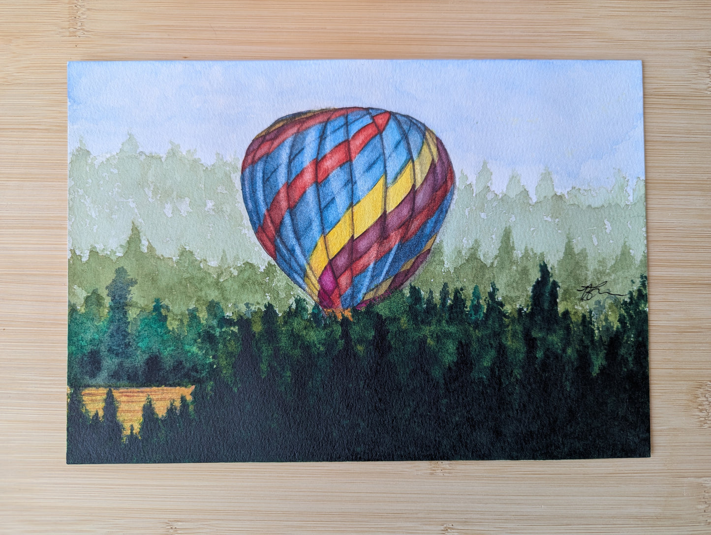 Painting of a hot air balloon taking off from a farmers field  resting on a wooden table. 