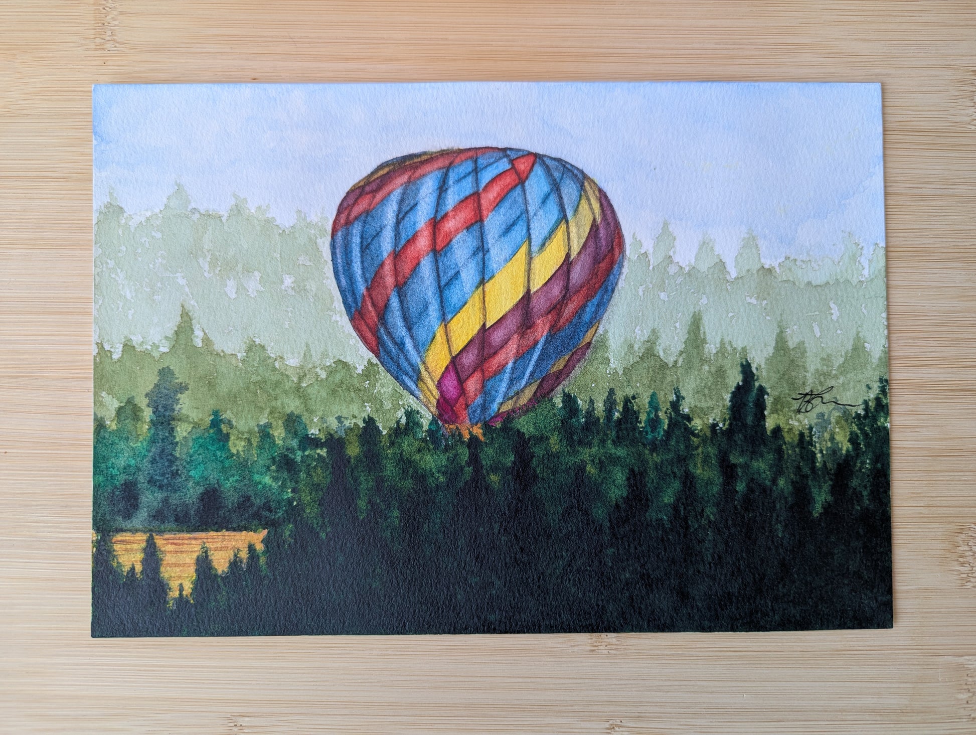 Painting of a hot air balloon taking off from a farmers field  resting on a wooden table. 