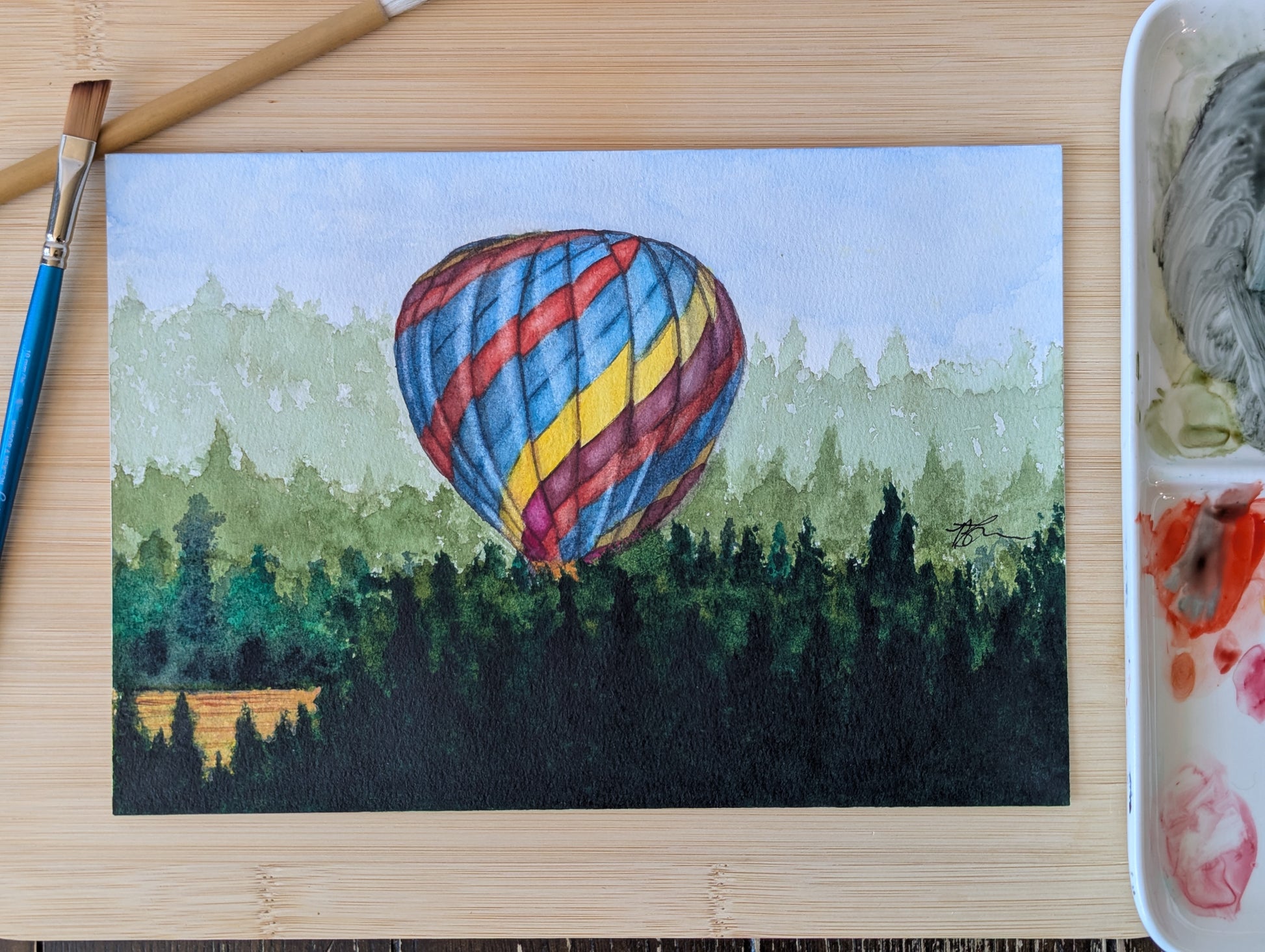 A painting of a colourful hot air balloon rests on a wooden table with a paint pallette and paintbrushes