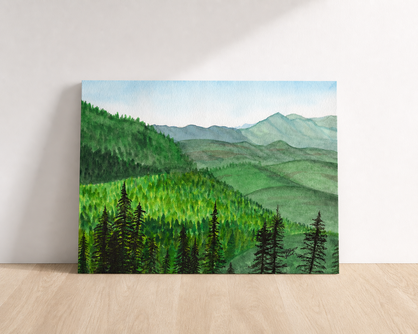 A painting of rolling green hills with pine trees in the foreround and bluish mountains in the background rests on a wood table and leans against a white wall