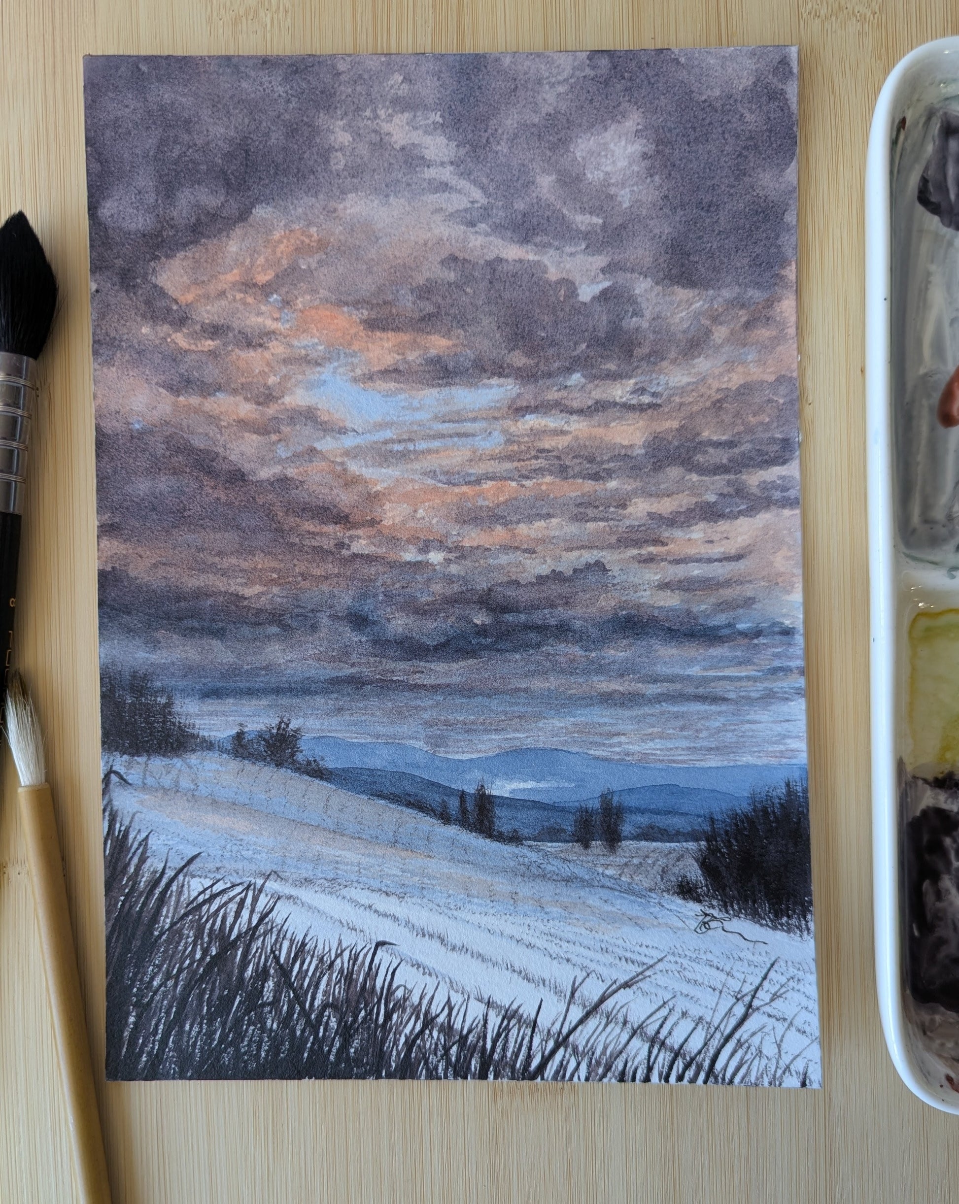 Watercolor Painting of a snowy landscape with a brush and paint palette on a wooden surface